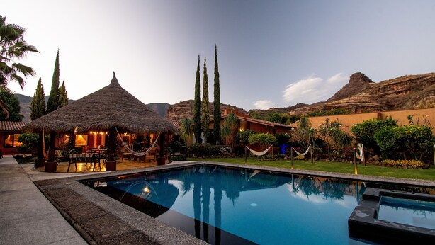 A pool area with a thatched-roof structure, palm trees, and a view of a mountainous landscape. - Quinta Ascension - Adults Only (Malinalco)