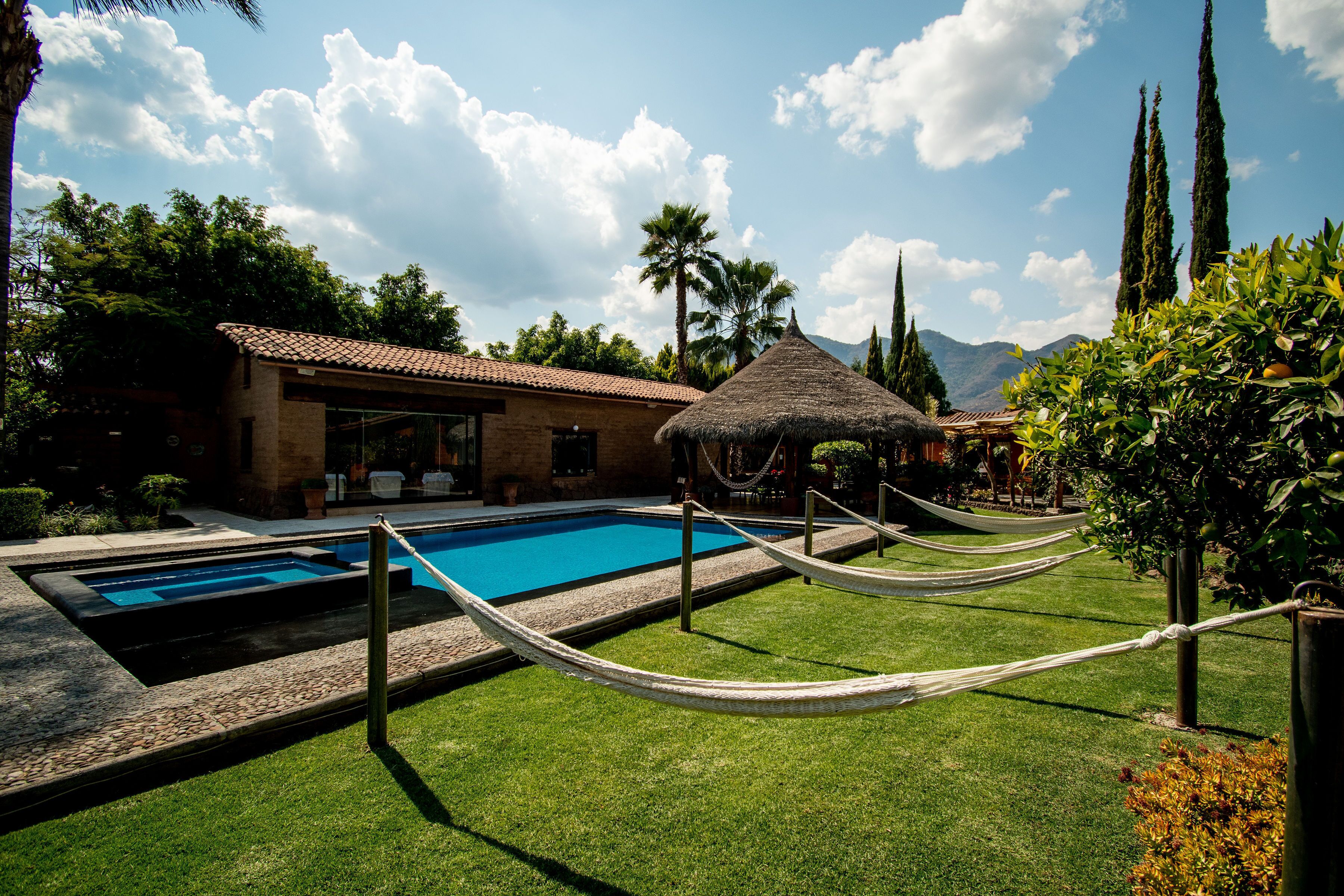 A swimming pool with a thatched-roof structure, surrounded by a grassy area and a building with a tiled roof.