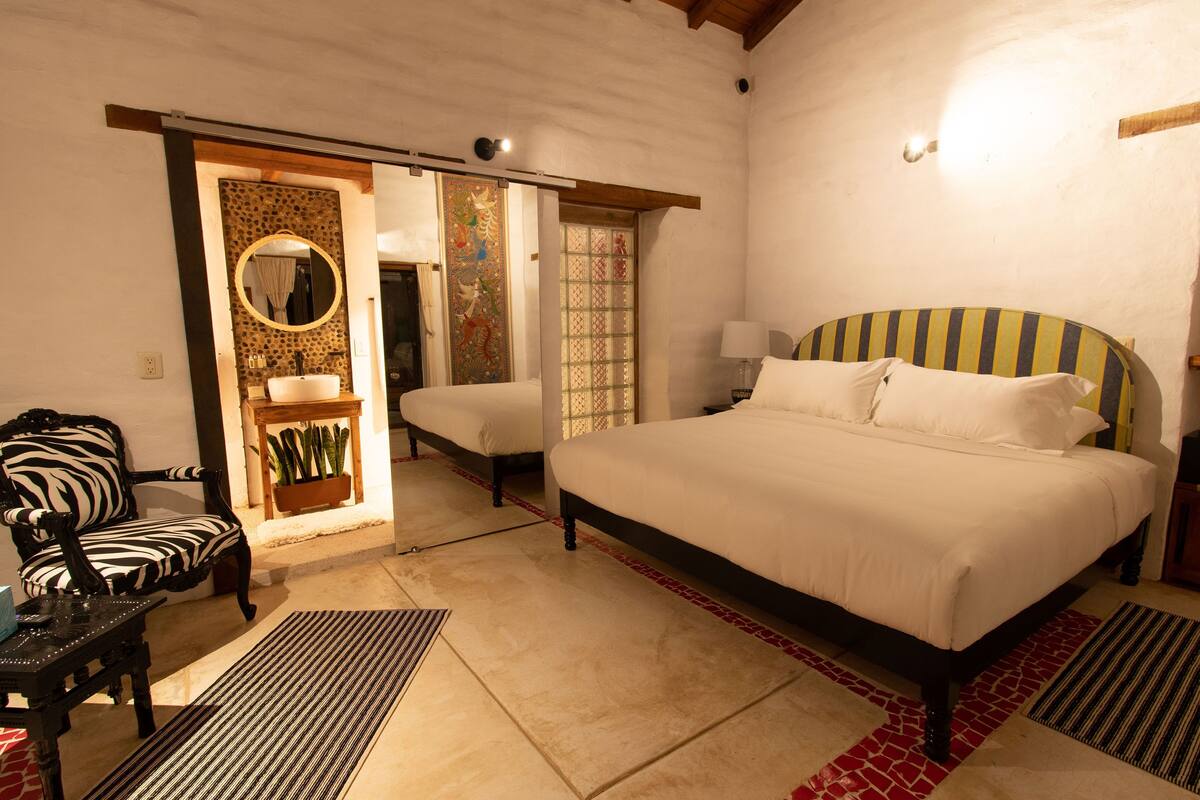 A bedroom with a large bed, a chair, a small table, and a mirror. There is a sliding door leading to another room.