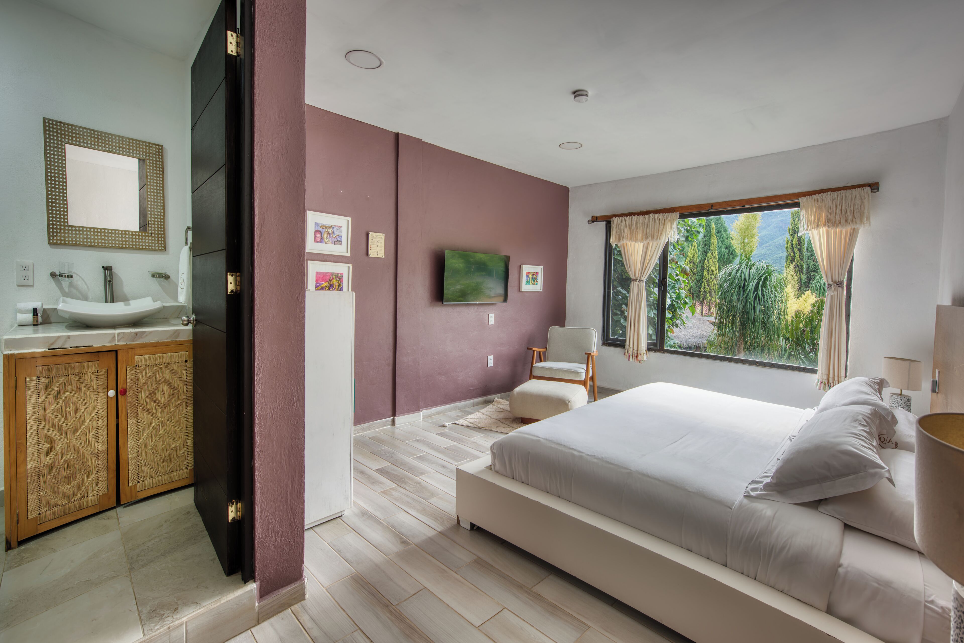 Un dormitorio moderno con una cama grande, un baño con lavamanos y espejo, y vistas a zonas verdes a través de la ventana.