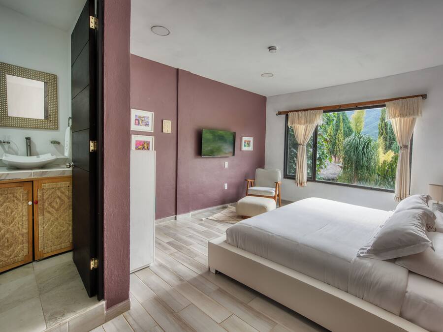 Un dormitorio moderno con una cama grande, un baño con lavamanos y espejo, y vistas a zonas verdes a través de la ventana.