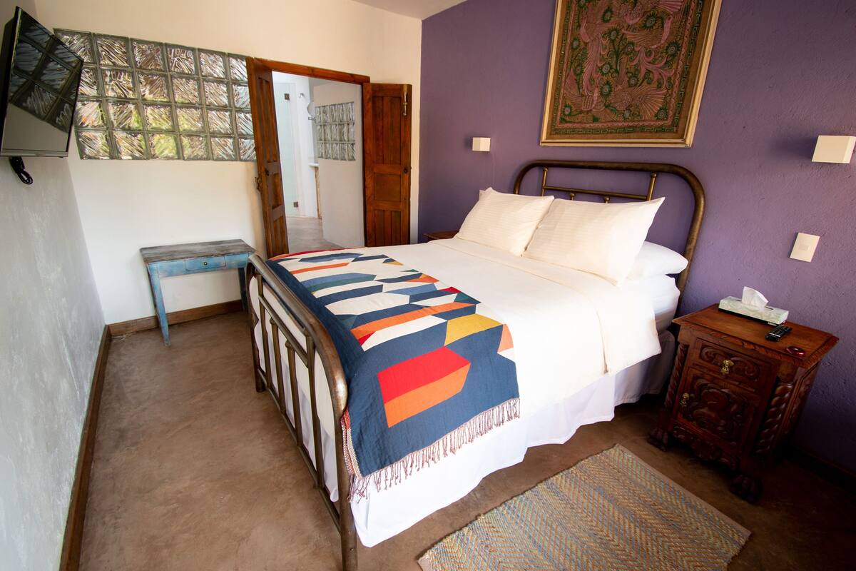 A bedroom with a bed, a wooden nightstand, a bench, a wall-mounted TV, and a door leading to another room.