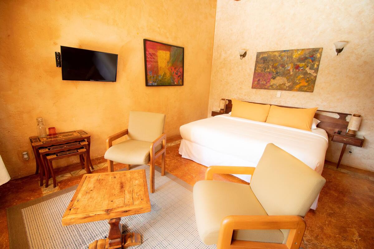 A hotel room with a bed, two chairs, a small table, a TV, and artwork on the walls.
