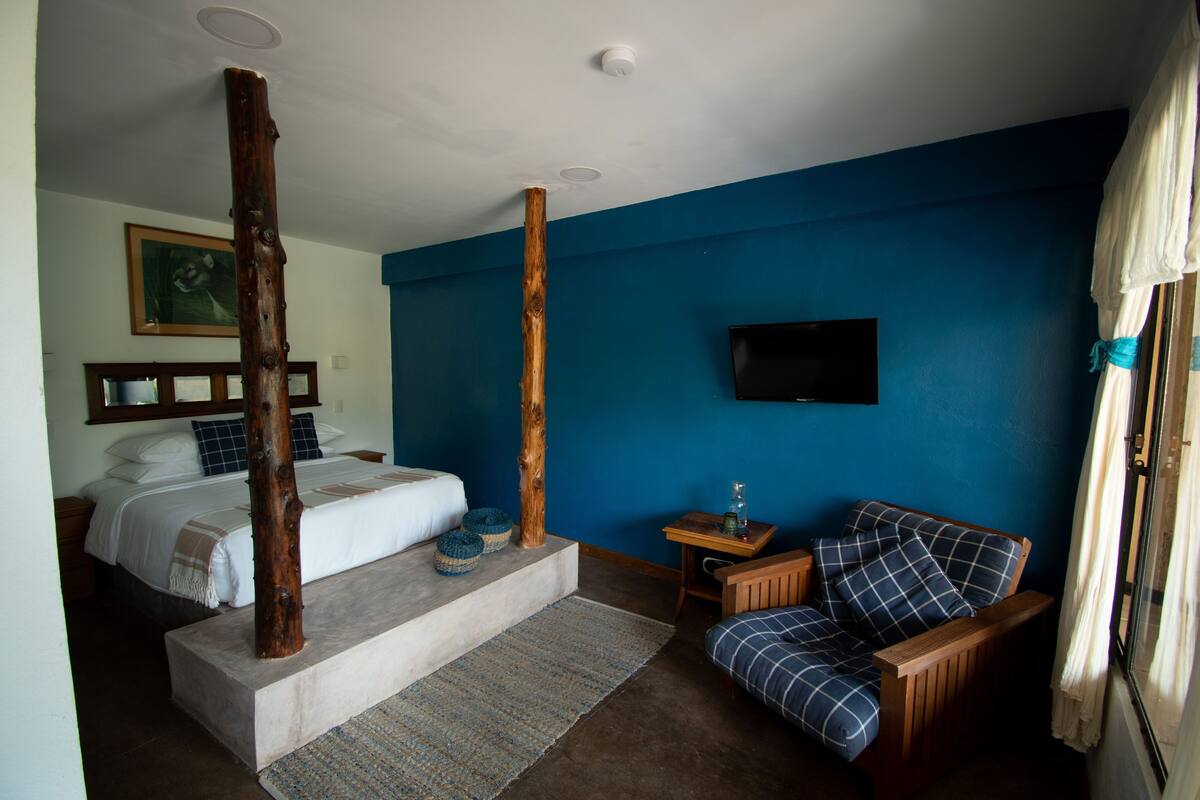 A bedroom with a bed, a wooden pillar, a blue wall, a television, a small table, and a window with curtains.