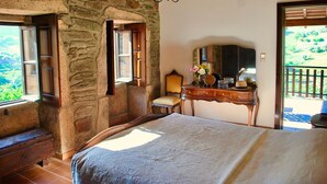 Une chambre avec un mur en pierre, un lit en bois, une commode avec un miroir et une fenêtre donnant sur un paysage verdoyant.