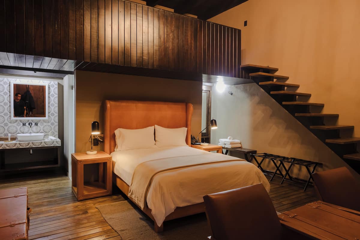 Ein Hotelzimmer mit einem hölzernen Bett, einem kleinen Nachttisch mit Lampe, einem Holzstuhl und einer Treppe, die zu einer oberen Ebene führt.