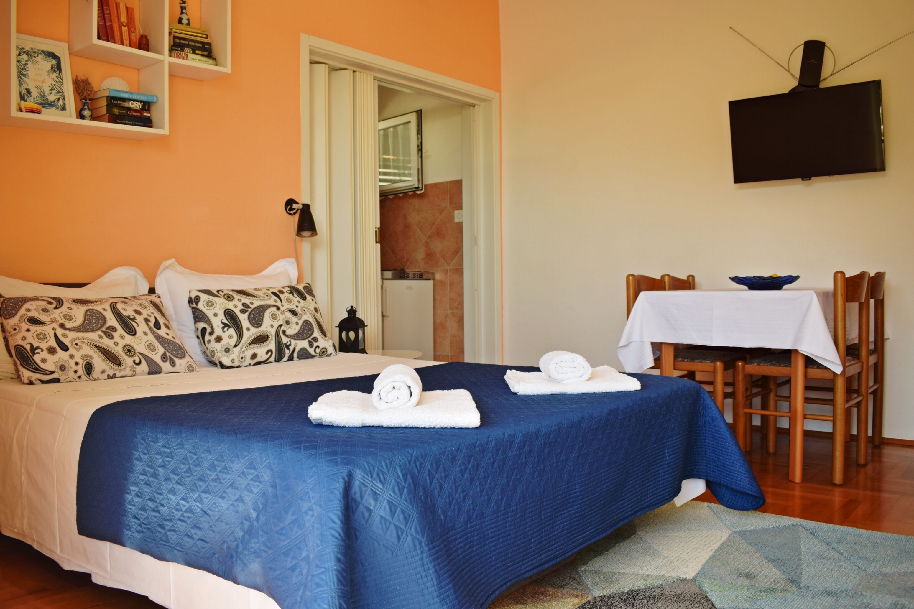 A neatly made bed with a blue bedspread, two pillows, and two rolled towels. There is a dining table with a white tablecloth and a bowl on it.