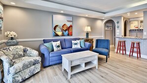 A living room with a blue sofa, a patterned armchair, a wooden coffee table, and a kitchen area with red stools.