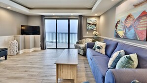 A modern living room with a large sofa, a wooden coffee table, and a TV. There is a balcony with a view of the sea.