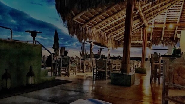 A beachside restaurant with outdoor seating, thatched roof structures, and a view of the ocean.