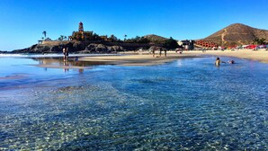 A beach with clear shallow waters, a large building with a tower, and a hill in the background.