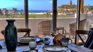 A dining table set with plates, a teapot, cups, and a vase. The table is in a room with wicker chairs and a view of the beach and a building in the distance.
