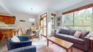 Un salon moderne avec un canapé, une table basse et des fauteuils, un coin repas avec une table et des chaises, une cuisine avec des meubles en bois et un décor aquatique, et de grandes fenêtres donnant sur la verdure.
