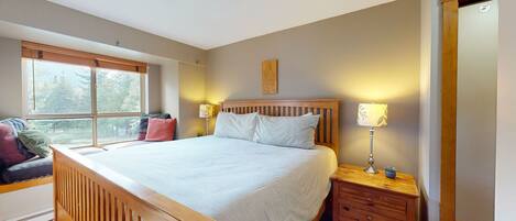 A bedroom with a large bed, a wooden nightstand with a lamp, a window with blinds, and a view of trees outside.