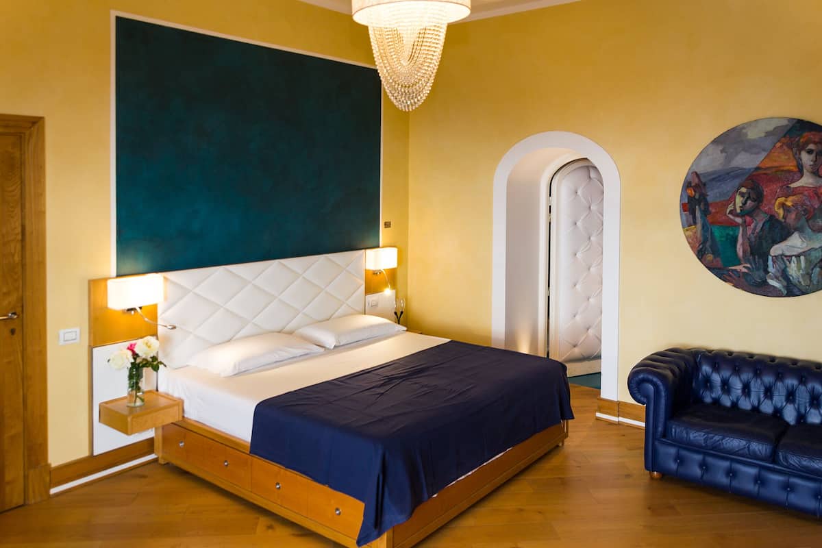 Un dormitorio con una cama grande, una mesita de noche con lámpara, un sofá azul y un cuadro en la pared.