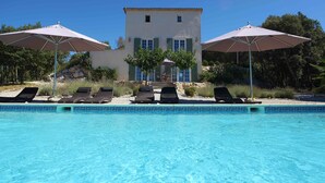 Una piscina con sillas de descanso y sombrillas, un edificio con contraventanas verdes y un cielo azul despejado.