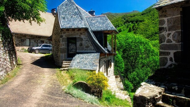 Ein Steinhaus mit einem steilen Dach, einem kleinen Balkon und einem Feldweg, der davorhin führt.