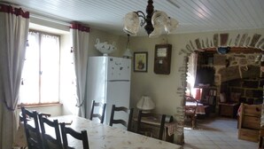 Ein Essbereich mit einem Tisch, der mit einer floralen Tischdecke bedeckt ist, umgeben von Holzstühlen. Ein weißer Kühlschrank steht an der Wand, und darüber an der Wand ist eine Uhr angebracht.