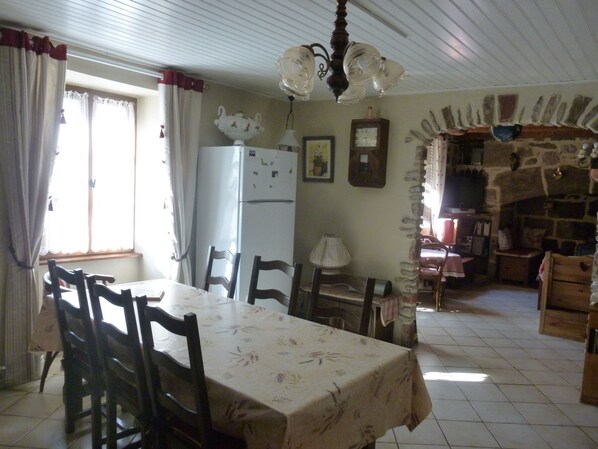 Un comedor con una mesa cubierta por un mantel floral, rodeada de sillas de madera. Un refrigerador blanco está apoyado contra la pared, y un reloj está colgado en la pared encima.