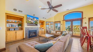 A spacious living room with a fireplace, a large sofa, a dining area, and a view of a residential area through large windows. - Semi-Oceanfront Premier Home w/ Pool, Hot Tub, Elevator, Game Room, and Theater (Avon)