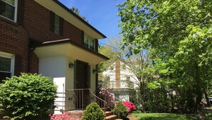 A two-story brick house with a front porch, a small garden with pink flowers, and a clear blue sky. - Unit2 GR8 LOCATION mins to Longwood Medical Area, BC, BU, HarvMedSchool SPACIOUS (Brookline)
