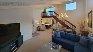 A spacious living room with a large TV, a blue sofa, and a glass coffee table. A staircase leads to an upper level with a dining area and kitchen. - Luv Oceanside-1 House to Beach (San Diego)