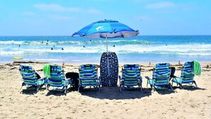 A beach with six striped beach chairs under a blue umbrella, a geometric patterned chair, and the ocean in the background. - Luv Oceanside-1 House to Beach (San Diego)