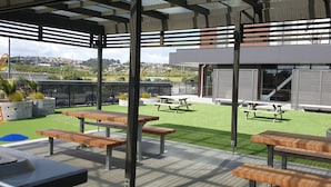 Un patio al aire libre con bancos de madera y mesas de picnic, rodeado por un edificio moderno con grandes ventanales y vistas a una zona residencial.