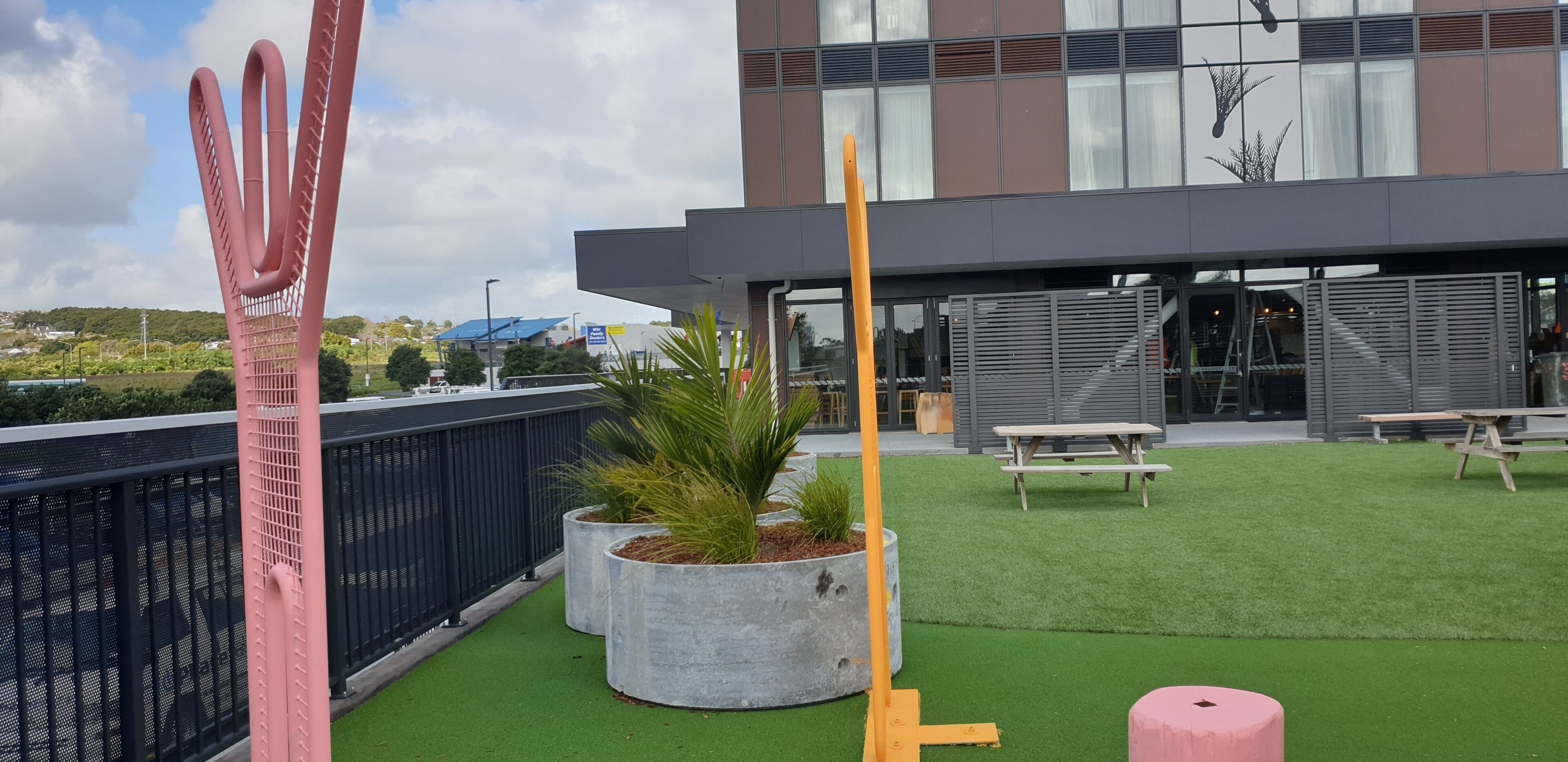 A modern building with a rooftop garden featuring artificial grass, concrete planters with plants, and outdoor seating.