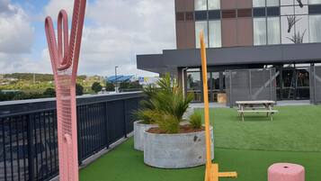 A modern building with a rooftop garden featuring artificial grass, concrete planters with plants, and outdoor seating.