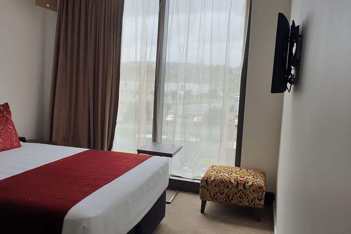 Ein Hotelzimmer mit Bett, einem Vorhangfenster, einem an der Wand montierten Fernseher und einem kleinen Tisch.