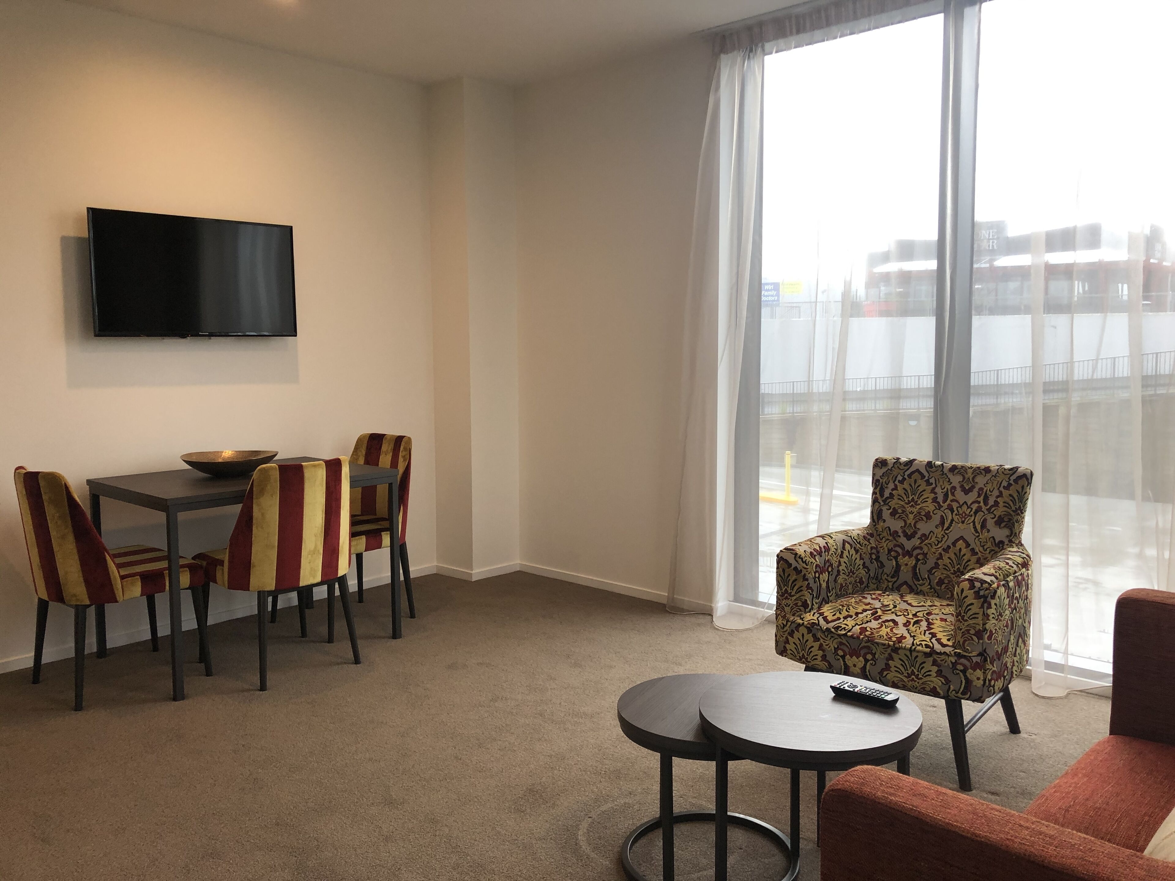 A hotel room with a flat-screen TV mounted on the wall, a dining table with chairs, a round coffee table, and a patterned armchair.