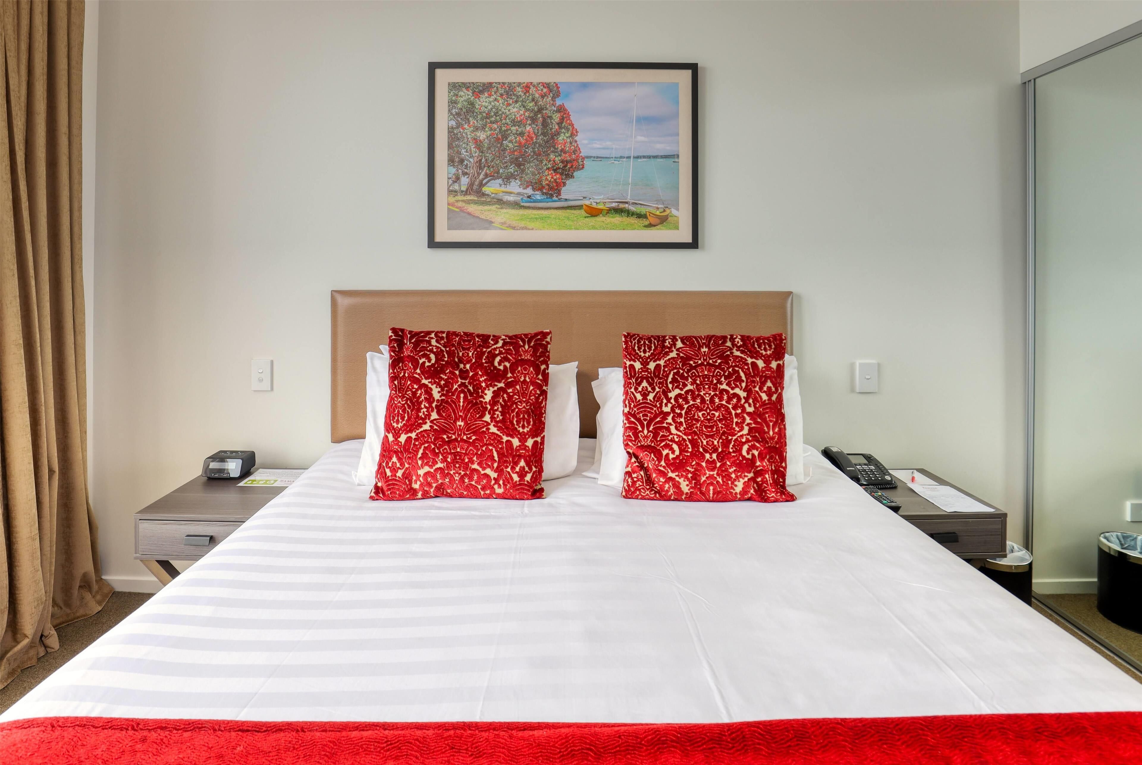 A hotel room with a large bed, two red patterned pillows, a wooden headboard, a painting of a tree and water on the wall, a bedside table with a phone and a lamp, and a window with curtains.
