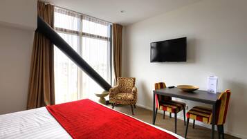 Quarto de hotel com cama, escrivaninha com cadeiras, televisão e janela com cortinas.