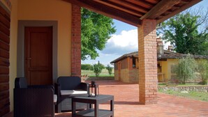 Un patio techado con mobiliario de mimbre y suelo de ladrillo, con vistas a un paisaje rural.