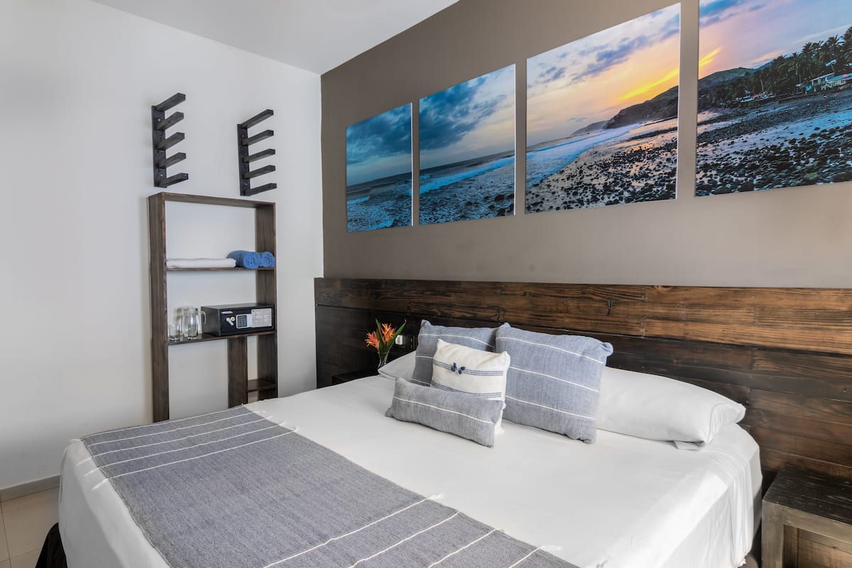 A bedroom with a bed, a wooden headboard, a shelf with towels, and a wall-mounted artwork of a beach scene.