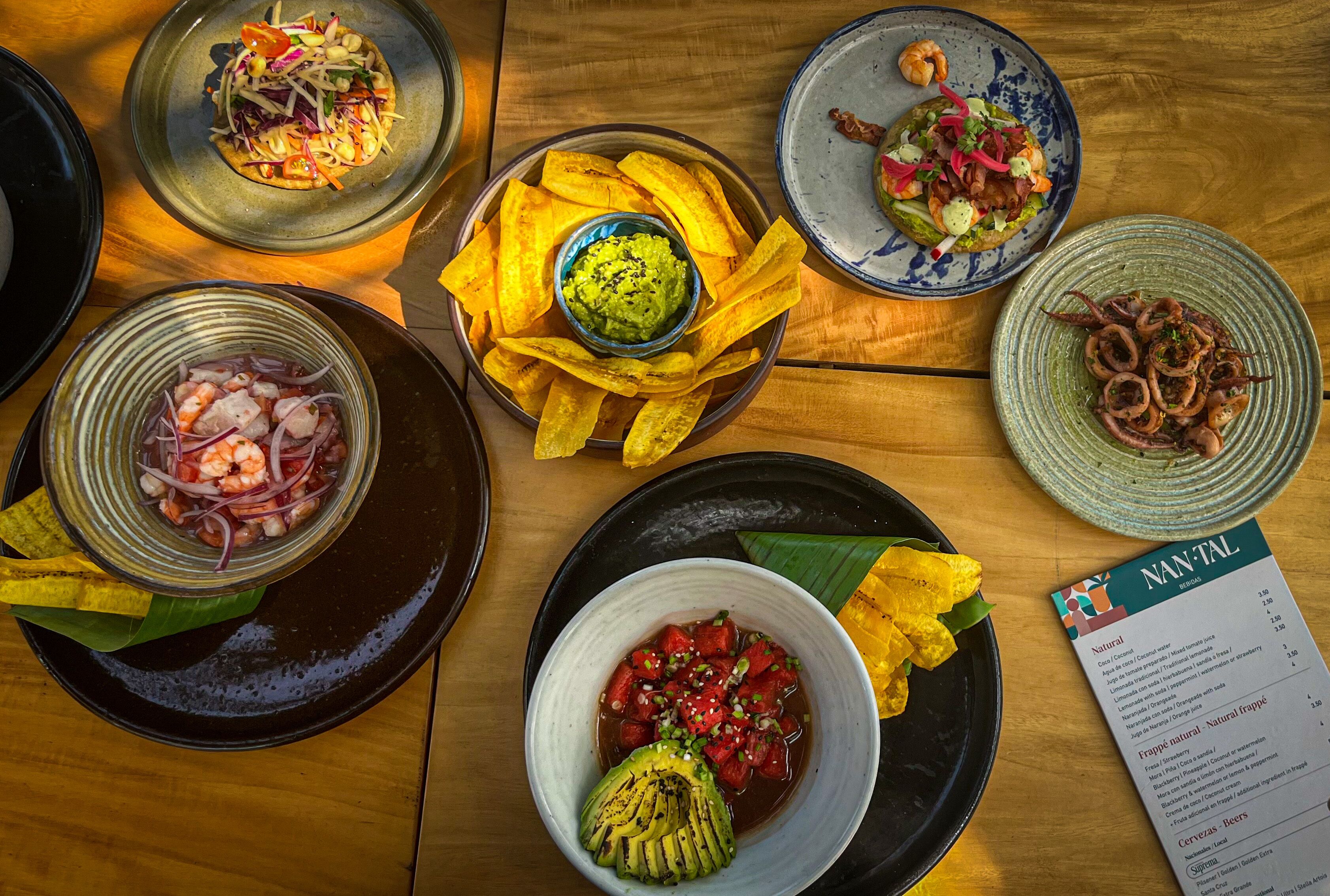 On peut voir une variété de plats sur une table en bois, notamment un saladier de salade aux crevettes, un bol de salsa à l’avocat et une assiette de bananes plantains frites.
