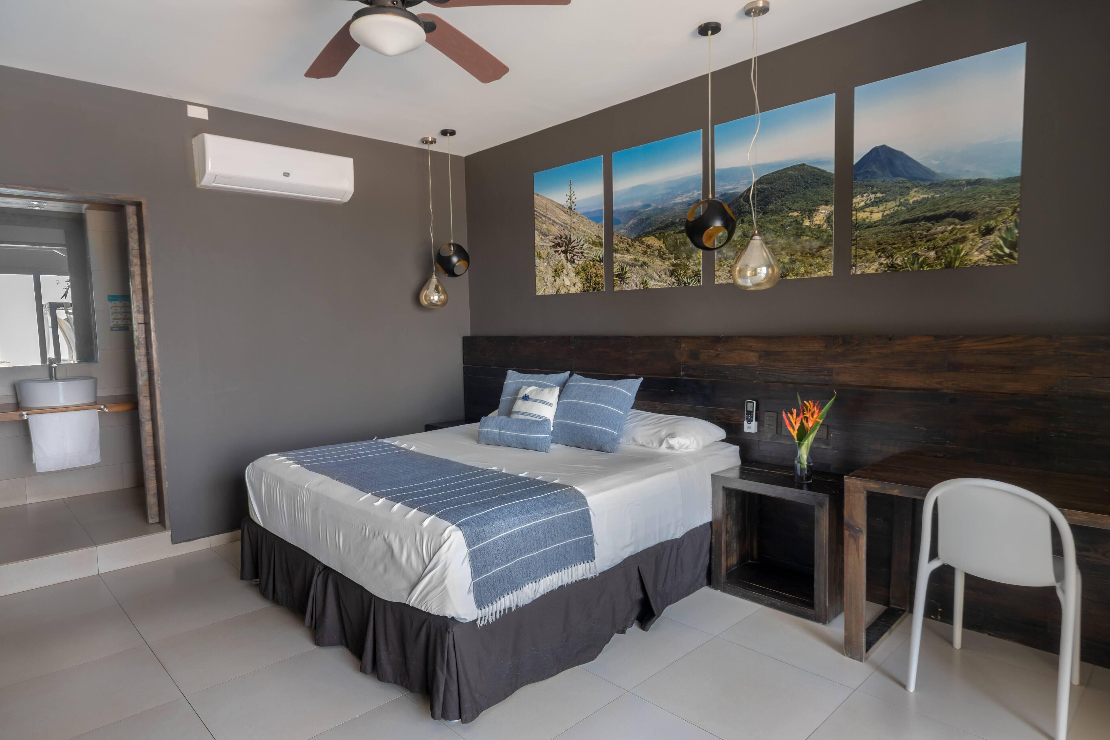 A modern bedroom with a large bed, a desk, and a chair. There are three wall-mounted picture frames and a ceiling fan.