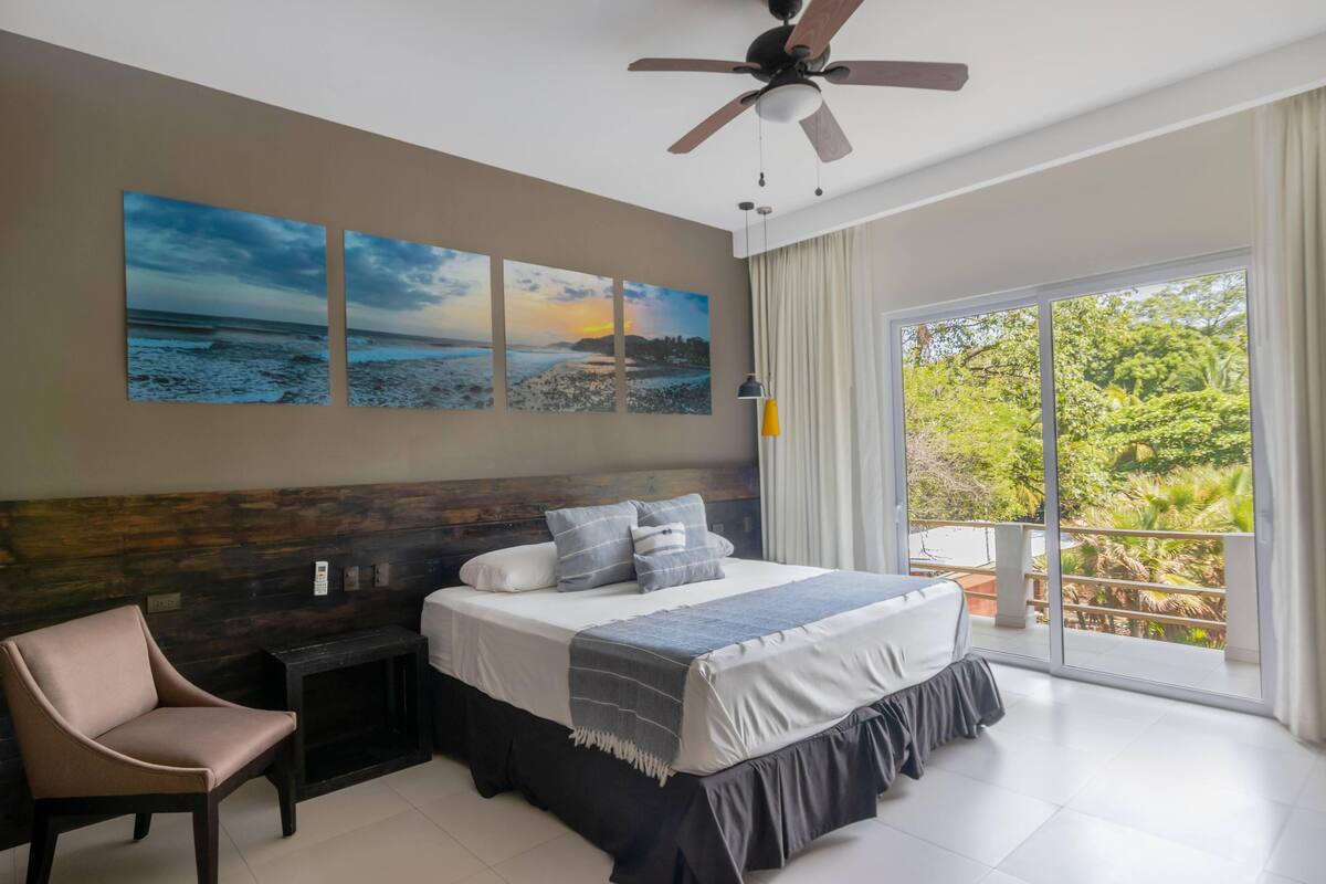 A bedroom with a bed, a chair, a nightstand, and a view of the outdoors.