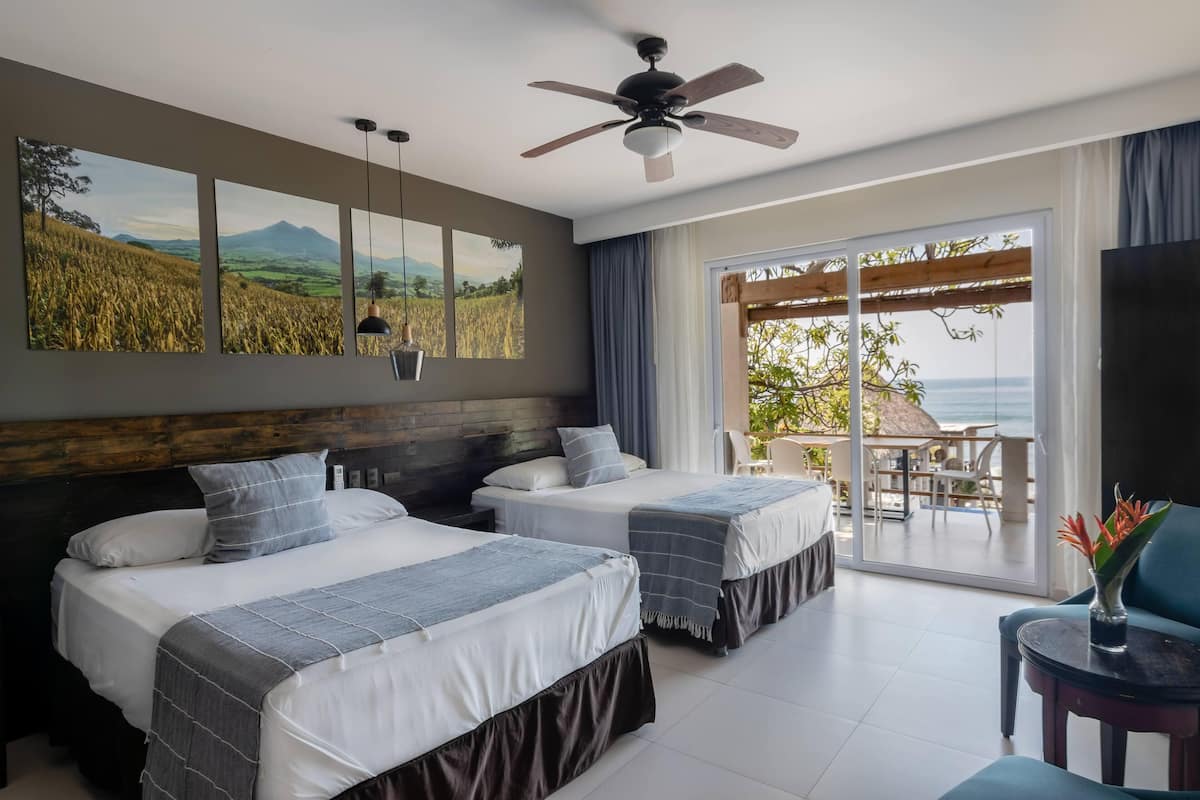 A hotel room with two beds, a ceiling fan, a large window with a view of the sea, and a balcony with outdoor furniture.