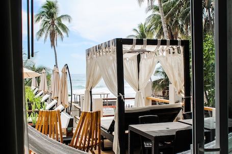 A beachside restaurant with outdoor seating, including wooden chairs and tables, and a view of the ocean.