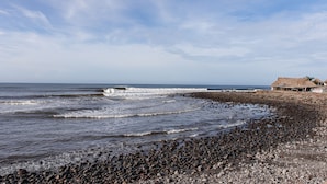 Una spiaggia rocciosa con onde, una costruzione con tetto di paglia e cielo sereno.