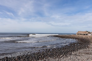 A rocky beach with waves, a thatched-roof structure, and a clear sky.