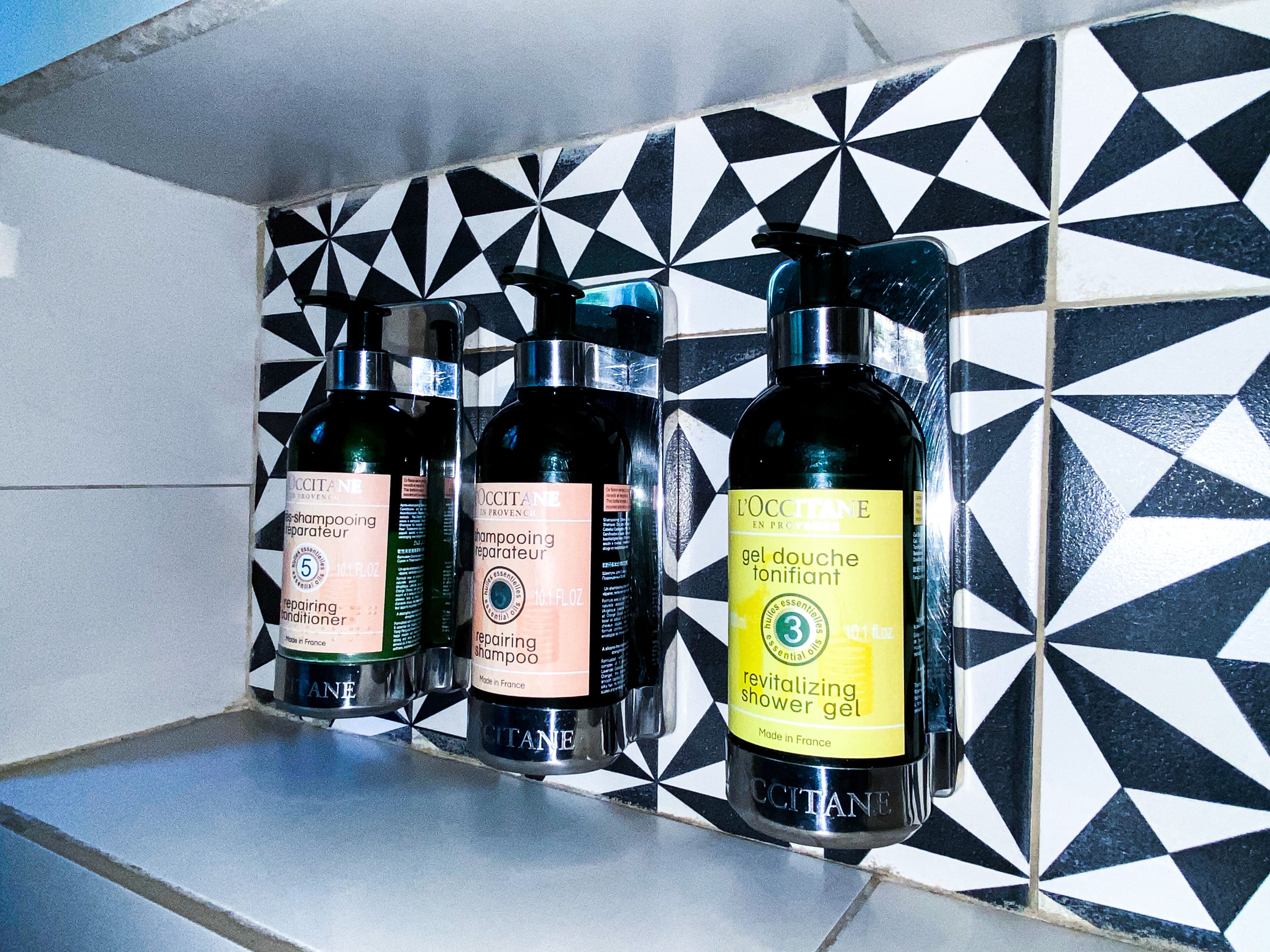 Three bottles of L'OCCITANE products on a tiled shelf.