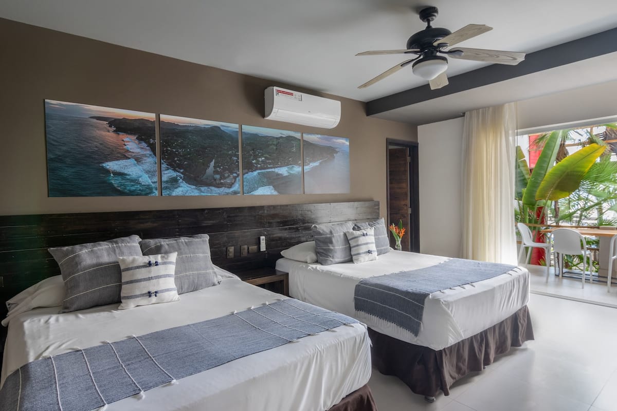 A hotel room with two beds, a ceiling fan, an air conditioning unit, and a large window with a view of the ocean.