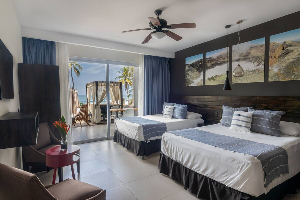 A hotel room with two beds, a ceiling fan, a TV, a small table with a vase of flowers, and a large window with a view of palm trees and the ocean.