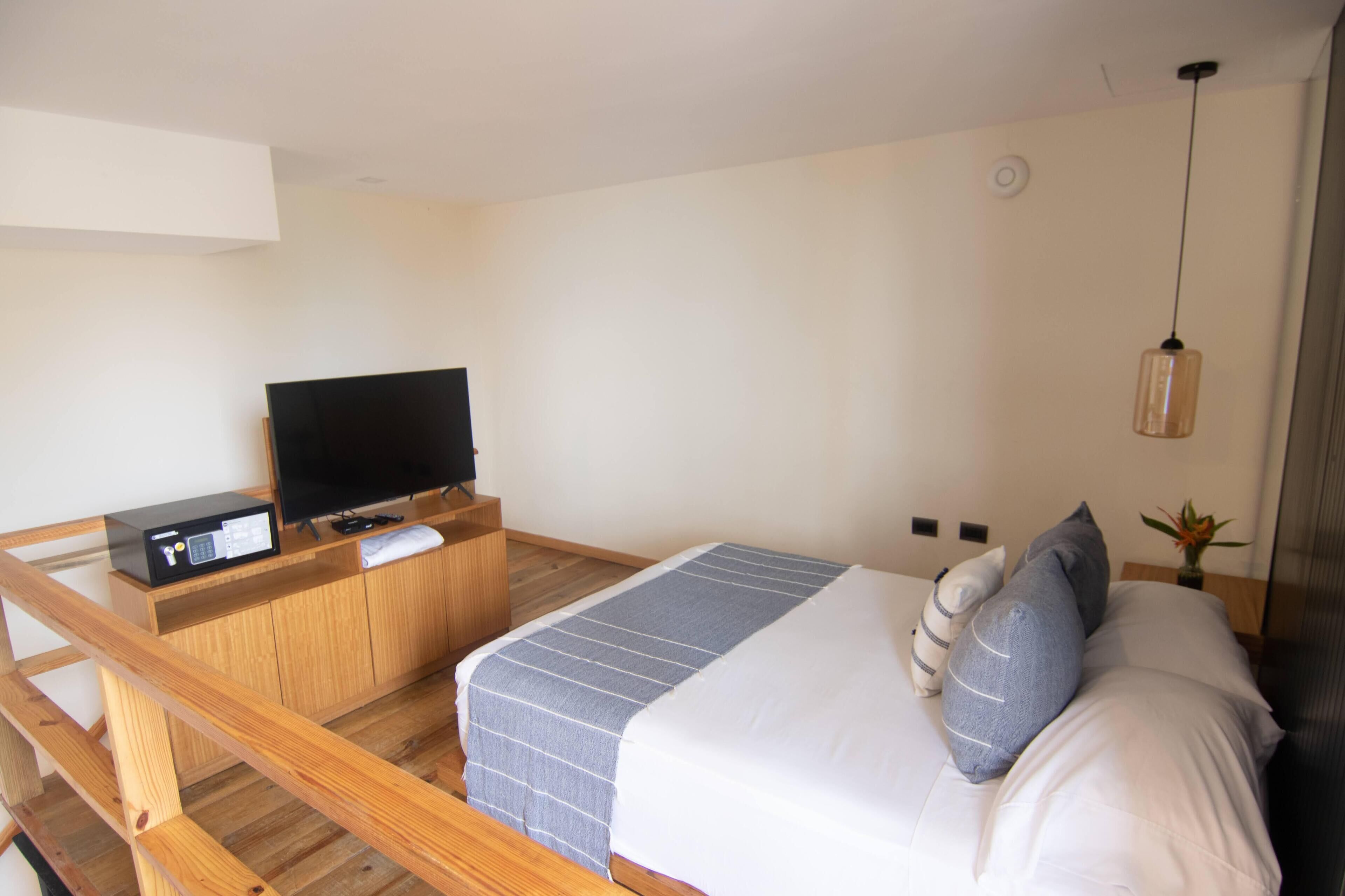 A hotel room with a bed, a TV on a wooden stand, a small table, and a hanging light fixture.