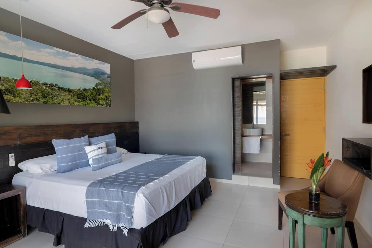 A bedroom with a bed, a ceiling fan, a painting, a bathroom, and a small table with a plant.
