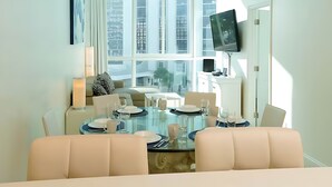 A modern dining area with a glass table, white chairs, and a city view through large windows. - NEW BEACHFRONT 2 bedroom CONDO-HOTEL with parking and direct beach access (Miami)