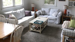 Ein gemütliches Wohnzimmer mit einer Sofagarnitur, einem Couchtisch und einem Esstisch mit Stühlen. Zwei Fenster mit Vorhängen, eine Lampe und Wandbilder sind ebenfalls vorhanden.